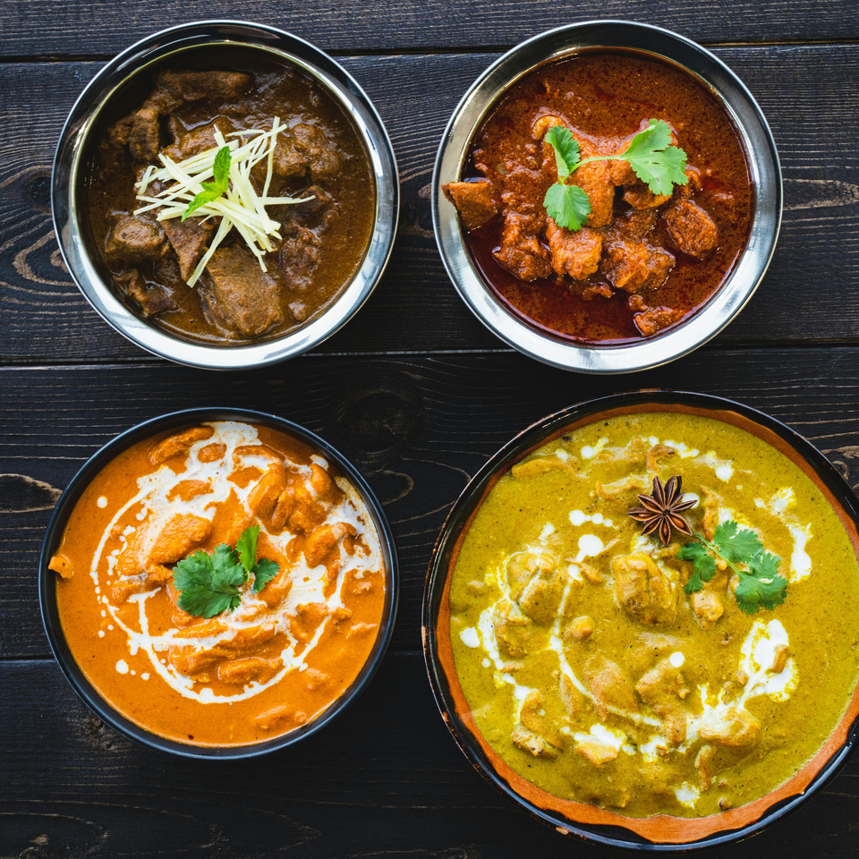 Four bowls of Indian curries, including butter chicken and green curry, on a dark wooden table.